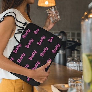Puede incluir: Una funda para port&aacute;til negra con un patr&oacute;n repetido de la palabra "Bratty" en letras rosas.
