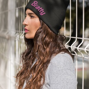 Puede incluir: Un gorro negro con la palabra "Bratty" bordada en rosa en la parte delantera.