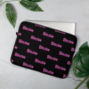 May include: Black laptop sleeve with the word "Bitchie" in pink repeated across the surface. The sleeve is rectangular and has a zipper closure. A laptop is partially visible inside the sleeve.