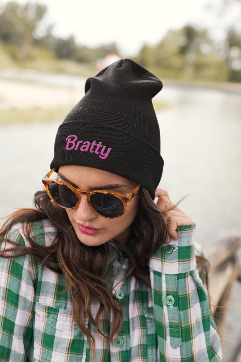 Puede incluir: Un gorro negro con la palabra "Bratty" bordada en rosa en la parte delantera. El gorro lo lleva puesto una persona con el pelo casta&ntilde;o y gafas de sol.