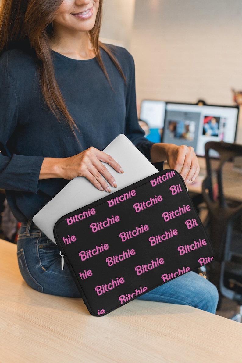 May include: Black laptop sleeve with the word "Bitchie" in pink repeated across the surface. The sleeve is held by a person, and a silver laptop is partially visible inside. The sleeve has a zipper closure.