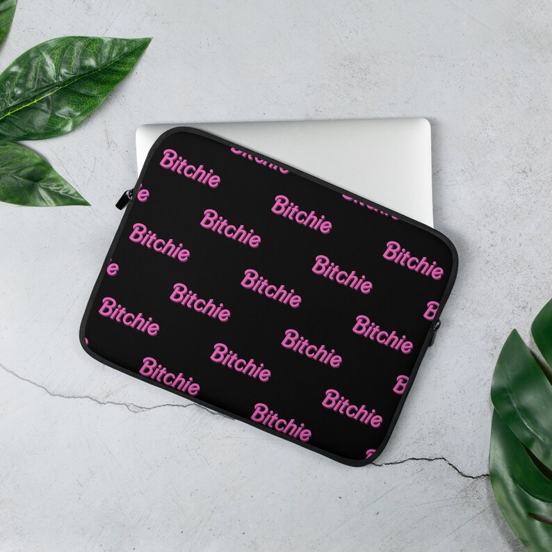 May include: Black laptop sleeve with the word "Bitchie" in pink repeated across the surface. The sleeve is on top of a silver laptop, with green leaves in the background.