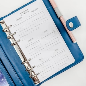 Peut inclure: Un agenda relié en cuir bleu avec des anneaux argentés et un stylo rose. L'agenda comporte un calendrier pour chaque mois de l'année, de janvier à juin. Le calendrier est imprimé sur du papier blanc.