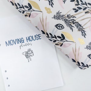 Może przedstawiać: Biały planer z okładką w czarno-białe wzory kwiatowe. Planer ma niebieski tytuł "Moving House Planner" i czarno-białą ilustrację osoby niosącej pudełka.