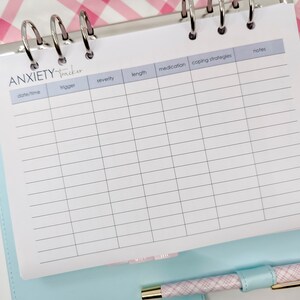 Pode incluir: Um rastreador de ansiedade branco com uma capa azul e rosa. O rastreador tem colunas para data/hora, gatilho, gravidade, duração, medicação, estratégias de enfrentamento e notas.