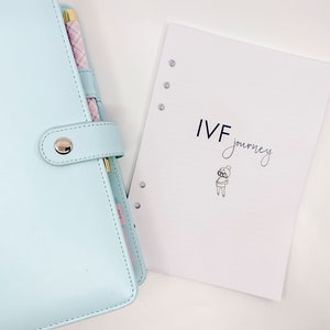 Peut inclure: Un agenda bleu clair avec un bouton argenté et un stylo qui sort du côté. L'agenda est ouvert sur une page avec le texte "IVF Journey" et un dessin simple d'une personne.