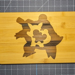 May include: Bamboo cutting board with a laser-etched design featuring a stylized cartoon character. The board has a natural wood grain finish and a stainless steel handle. The design is a darker shade of brown.