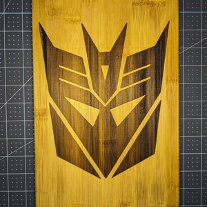 May include: A rectangular bamboo cutting board with a dark brown, stylized emblem burned into the wood. The board has a silver metal handle and is set against a dark background. The emblem is a geometric design.