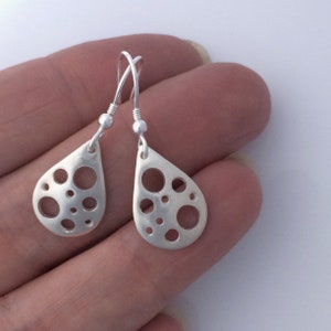 May include: A pair of silver teardrop-shaped earrings with a cutout design of circles. The earrings are hanging from a person's hand.