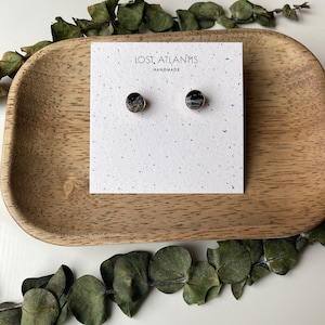 May include: A pair of round stud earrings with a gold-colored border. The earrings have a dark, speckled center. The earrings are displayed on a white card with the text "LOST ATLANTIS HANDMADE". The card is on a wooden tray.