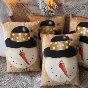 May include: Four tan-colored throw pillows with a snowman design. Each pillow has a black hat with a yellow and white polka dot band. The snowman's face is painted with black eyes, a red nose, and a black mouth. The pillows have the word "Jingles" written on them.
