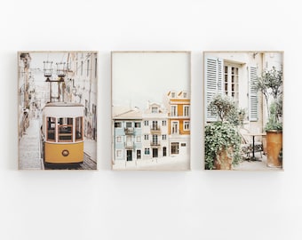 May include: Three framed photographs of a city street scene. The first photo shows a yellow tram traveling down a cobblestone street lined with buildings. The second photo shows a row of colorful buildings with a blue sky in the background. The third photo shows a cafe patio with a table and chairs, potted plants, and a white building with a window.
