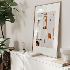 Op de afbeelding: Een ingelijste foto van een wit gebouw met oranje accenten, hangend aan een witte muur boven een witte commode met een witte kom en een boek erop.