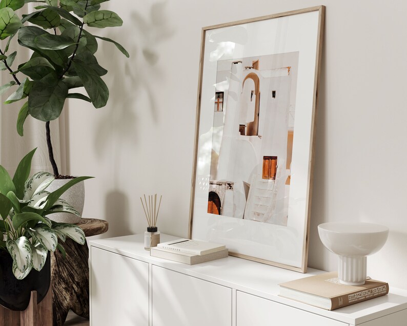Op de afbeelding: Een ingelijste foto van een wit gebouw met oranje accenten, hangend aan een witte muur boven een witte commode met een witte kom en een boek erop.