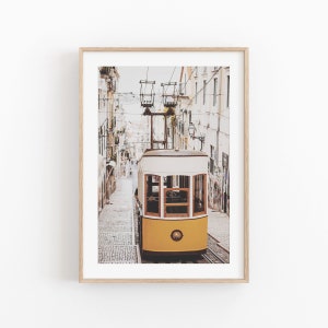 May include: Framed print of a yellow and white vintage tram descending a cobblestone street in Lisbon, Portugal. The tram is the focal point, set against a backdrop of buildings and overhead power lines. The print is in a light wood frame.