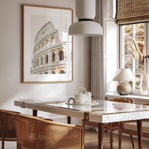 May include: A dining room with a white table with a white and gold inlay, two wicker chairs, a white ceramic teapot and a white pendant light hanging over the table. A framed print of the Colosseum in Rome hangs on the wall behind the table.
