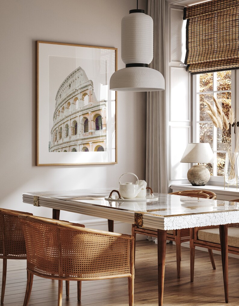 May include: A dining room with a white table with a white and gold inlay, two wicker chairs, a white ceramic teapot and a white pendant light hanging over the table. A framed print of the Colosseum in Rome hangs on the wall behind the table.