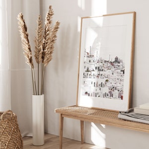 May include: A framed print of a white cityscape with a light blue sky. The print is hanging on a white wall next to a vase with dried pampas grass.