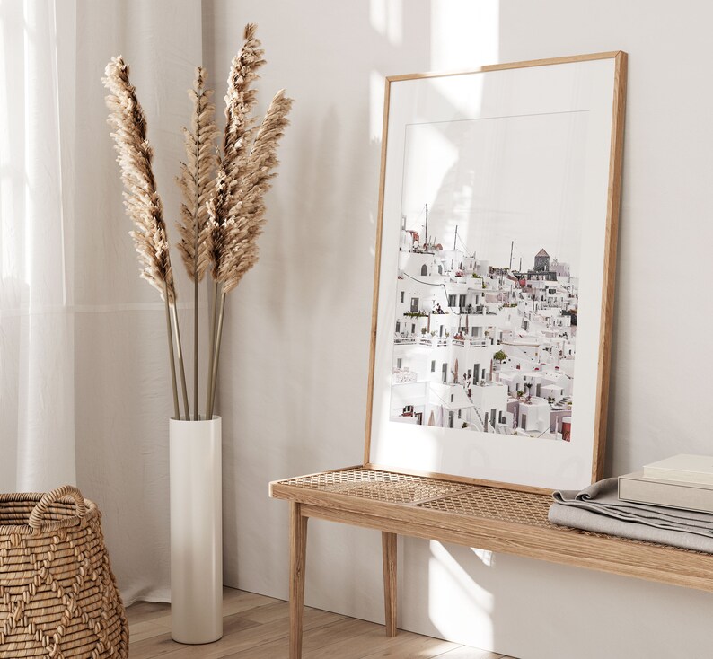 May include: A framed print of a white cityscape with a light blue sky. The print is hanging on a white wall next to a vase with dried pampas grass.
