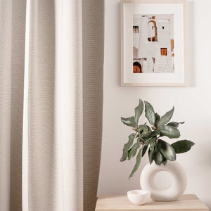 Op de afbeelding: Een lichtbeige linnen gordijn hangt naast een houten nachtkastje met een geweven lade. Een witte keramische vaas met een ronde opening bevat een groene, bladerrijke plant. Een ingelijste foto van een wit gebouw met een deur en treden hangt aan de muur boven het nachtkastje.