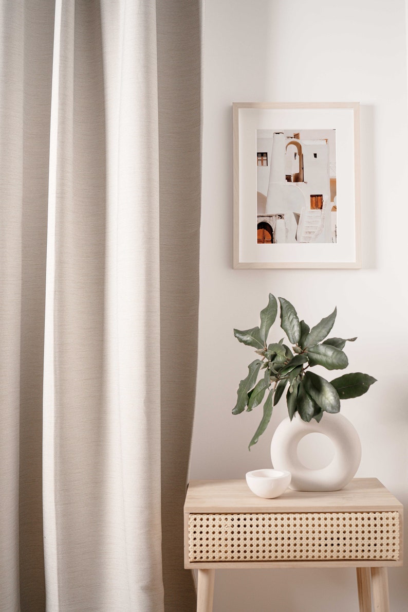Op de afbeelding: Een lichtbeige linnen gordijn hangt naast een houten nachtkastje met een geweven lade. Een witte keramische vaas met een ronde opening bevat een groene, bladerrijke plant. Een ingelijste foto van een wit gebouw met een deur en treden hangt aan de muur boven het nachtkastje.