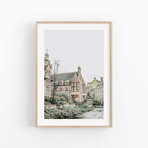 May include: A framed print showing a historic stone building with a red-tiled roof and multiple windows. The building is surrounded by greenery and sits next to a river. The print is in a light wood frame.
