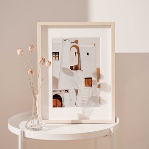 Op de afbeelding: Een ingelijste foto van een wit gebouw met een boog en een houten deur. Het gebouw bevindt zich op een zonnige locatie en heeft een witgekalkte buitenkant. De foto staat op een wit bijzettafeltje met een vaas met gedroogde bloemen.