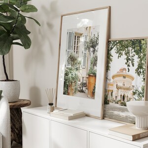 May include: Two framed photographs on a white shelf. The left photo shows a Parisian courtyard with a potted plant and a table and chairs. The right photo shows a building with a dome and a lush green tree.