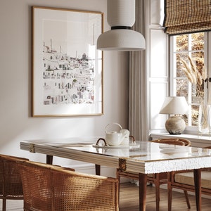 May include: A dining room with a white table with a white and gray mosaic top, four woven cane chairs, a white ceramic pendant light, and a framed print of a white cityscape.