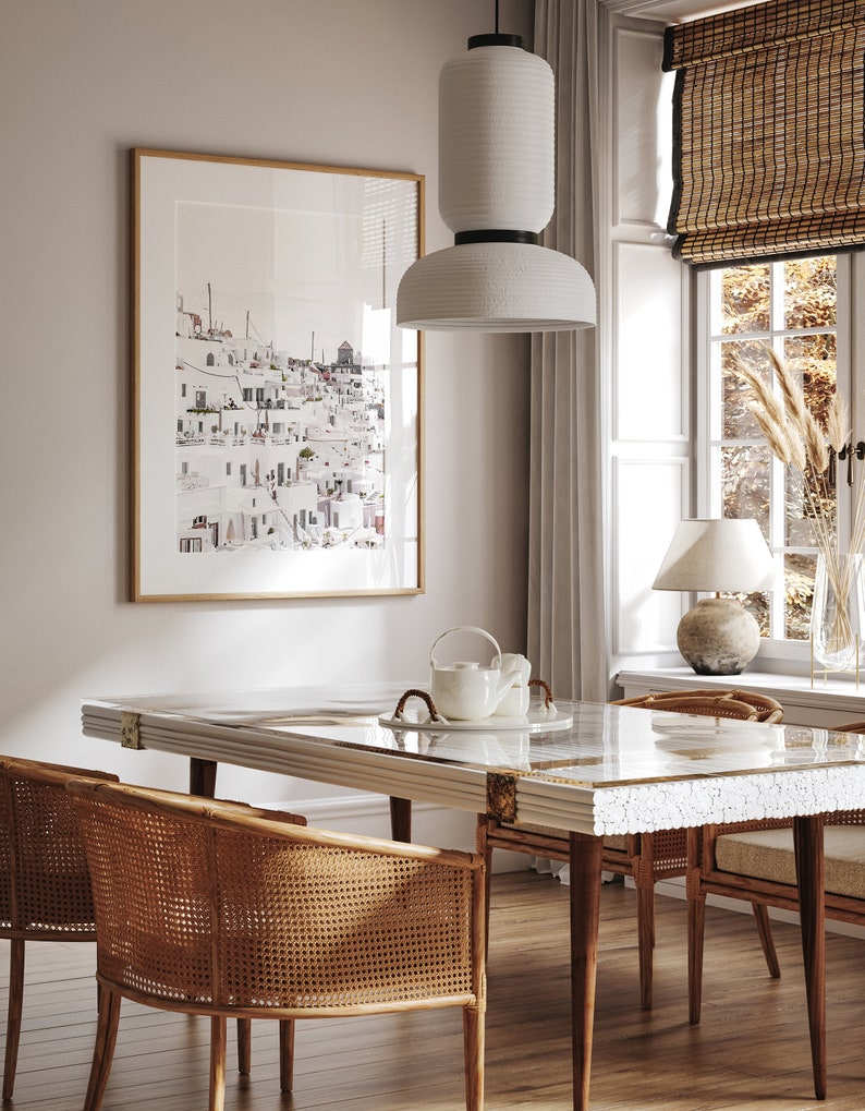 May include: A dining room with a white table with a white and gray mosaic top, four woven cane chairs, a white ceramic pendant light, and a framed print of a white cityscape.