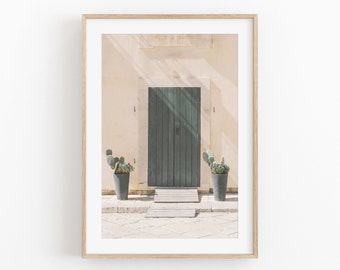 May include: A framed photograph of a green wooden door with two potted cactus plants in front of it. The door is set in a white wall with a light brown wooden frame. The door is slightly open and there are steps leading up to it.