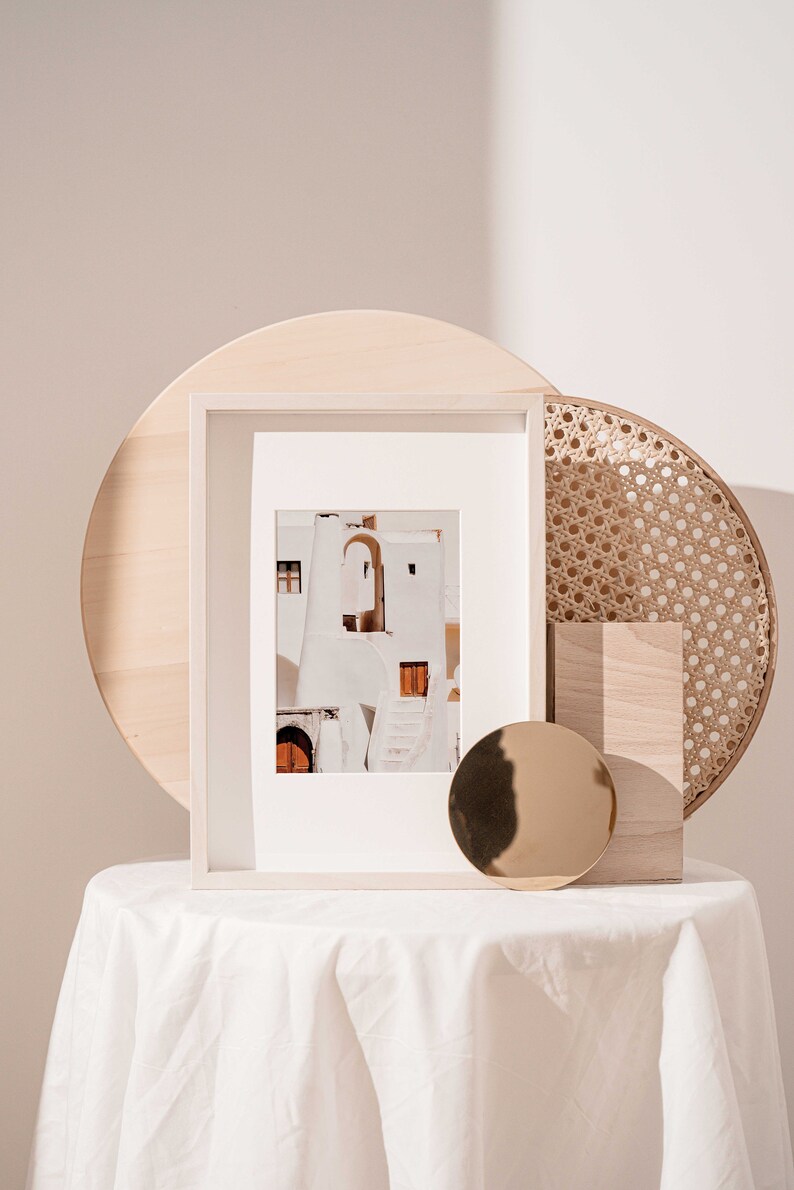 Op de afbeelding: Een wit ingelijste foto van een wit gebouw met een boog en een bruine deur. De foto staat op een witte tafel met een ronde houten dienblad, een geweven dienblad en een rond gouden object.