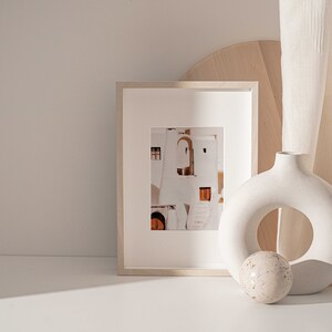 Op de afbeelding: Een witte keramische vaas met een ronde opening in het midden, een wit ingelijste foto van een wit gebouw met oranje deuren en een rond wit en bruin decoratief object op een wit oppervlak.