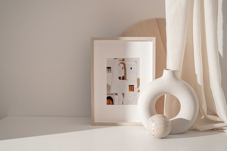 Op de afbeelding: Een witte keramische vaas met een ronde opening in het midden, een wit ingelijste foto van een wit gebouw met oranje deuren en een rond wit en bruin decoratief object op een wit oppervlak.