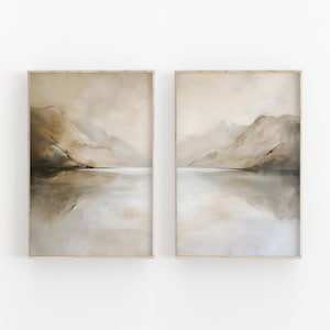 以下が含まれることがあります： 額縁に入った抽象的な風景画2枚セット。絵画は、茶色、ベージュ、灰色など、落ち着いた色合いで描かれています。絵画は、静かな水面に映る山脈を描いています。
