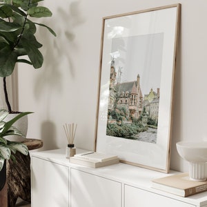 May include: A framed print depicting a European town with buildings and a river. The print is displayed on a white cabinet, alongside a plant, books, and a decorative bowl. The frame is a light wood colour.
