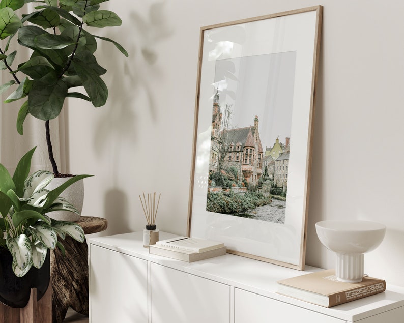 May include: A framed print depicting a European town with buildings and a river. The print is displayed on a white cabinet, alongside a plant, books, and a decorative bowl. The frame is a light wood colour.