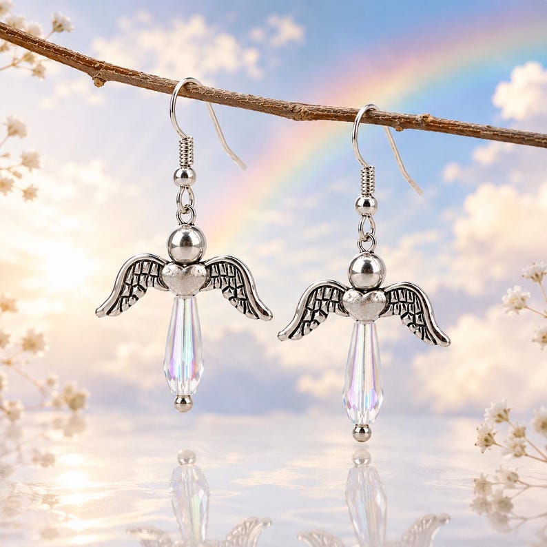 Op de afbeelding: Een paar zilverkleurige oorbellen met engelvleugels en iriserende kristallen traanvormige hangers. De oorbellen hangen aan een tak tegen een achtergrond van een regenboog en een bewolkte lucht. Een delicaat accessoire.