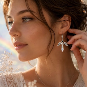 Op de afbeelding: Een vrouw draagt een zilveren engelenvleugeloorbel met een helder kristallen lichaam. De oorbel is opgehangen aan een zilveren haak. De vrouw draagt een witte kanten top en een zilveren ketting. Een regenboog is zichtbaar op de achtergrond.