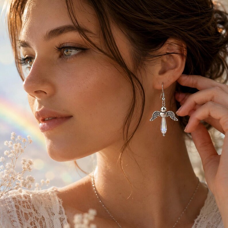 Op de afbeelding: Een vrouw draagt een zilveren engelenvleugeloorbel met een helder kristallen lichaam. De oorbel is opgehangen aan een zilveren haak. De vrouw draagt een witte kanten top en een zilveren ketting. Een regenboog is zichtbaar op de achtergrond.