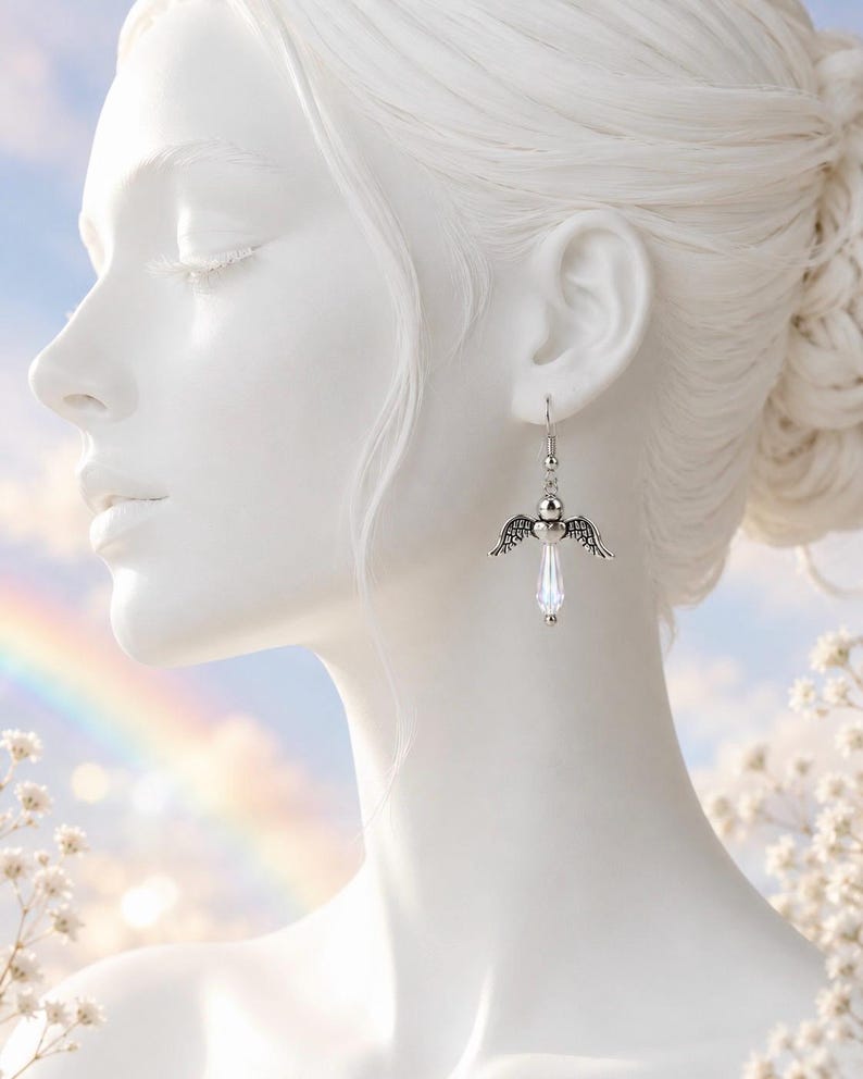 Op de afbeelding: Zilveren engelenvleugel oorbellen met een heldere kristallen druppelhanger. De oorbellen hangen aan een eenvoudige haak en hebben een kleine zilveren bal boven de vleugels. De achtergrond bevat een regenboog en zachte, vage kleuren.