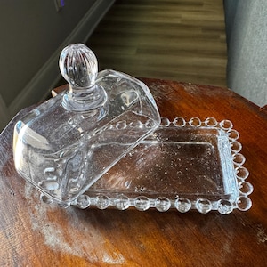 May include: A clear glass butter dish with a lid. The rectangular dish has a beaded edge and a domed lid with a round, ribbed handle. The dish is sitting on a wooden surface.