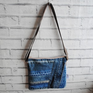 May include: A denim crossbody bag with a brown strap hangs against a white brick wall. The bag is made from various shades of blue denim, with a zip closure and a light-coloured lining. The strap is adjustable.