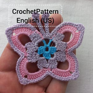 Pode incluir: Uma borboleta de crochê feita à mão em tons de rosa, lavanda e azul. A borboleta tem um design detalhado e vazado com fio metálico prateado. O texto "Crochet Pattern English (US)" é visível.