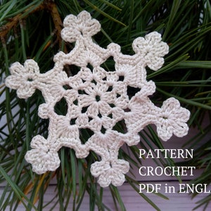 Könnte beinhalten: Ein weißer gehäkelter Schneeflocken-Anhänger mit einem filigranen Muster. Die Schneeflocke liegt auf einem grünen Tannenzweig. Der Text "PATTERN CROCHET PDF in ENGLISH" ist unterhalb der Schneeflocke sichtbar.