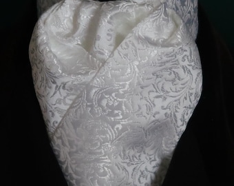 May include: A formal white ascot and matching neckwear set. The fabric features a subtle, ornate floral pattern. The ascot is folded and tied, with a matching flower accent. The set is designed for formal occasions.
