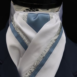 May include: A white and blue patterned stock tie with a silver lace trim. The tie is folded and displayed on a black mannequin.