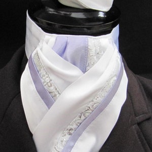 Puede incluir: Una corbata de montar a caballo blanca y lavanda con ribete de encaje plateado. La corbata está doblada y se muestra sobre un fondo negro.