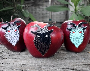 May include: Three red apples with black, white, and blue goat head pins on them. The goat heads have horns, a crown of thorns, and red eyes.