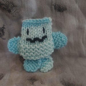 May include: A small, light blue knitted plush toy with a textured, garter-stitch body. The figure features two small arms extending from the sides, two rounded feet at the base, and a simple embroidered face with two black eyes and a wavy mouth.